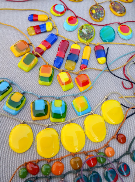 A close-up of multiple colorful glass pendant necklaces. The necklaces feature glossy, translucent rectangular, oval, and round pendants in bright yellow, green, blue, red, orange, and some with multicolor marbled patterns.