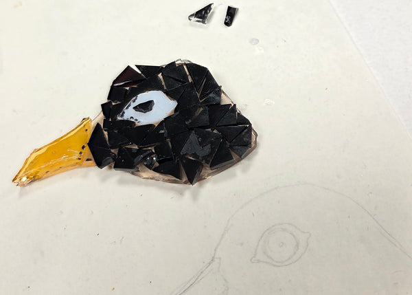 A close-up of a mosaic bird head craft piece made from black, white, and yellow glass fragments, placed on a white surface next to a clear container filled with colorful broken glass pieces.