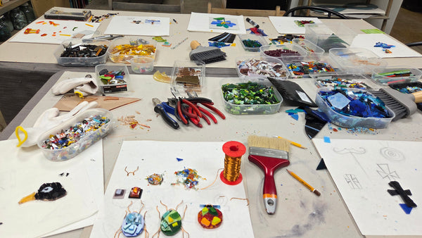 A cluttered craft table filled with colorful glass shavings, beads, wire, pliers, a paintbrush, and small glass art pieces arranged on white sheets of paper.