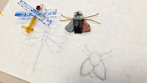A handmade stained glass insect craft, resembling a beetle or moth, displayed on a white sheet of paper next to pencil sketches of dragonflies and a moth. The craft features a blue and white patterned glass body, a brown and black patterned wing, and gold-toned metal legs and antennae.